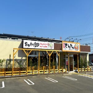 黄色い外壁の平屋の店舗。「おかわり自由」の看板と「とんかつ よし平」の看板が掲げられ、手前に白線を引いた駐車場がある