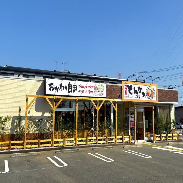 黄色い外壁の平屋の店舗。「おかわり自由」の看板と「とんかつ よし平」の看板が掲げられ、手前に白線を引いた駐車場がある