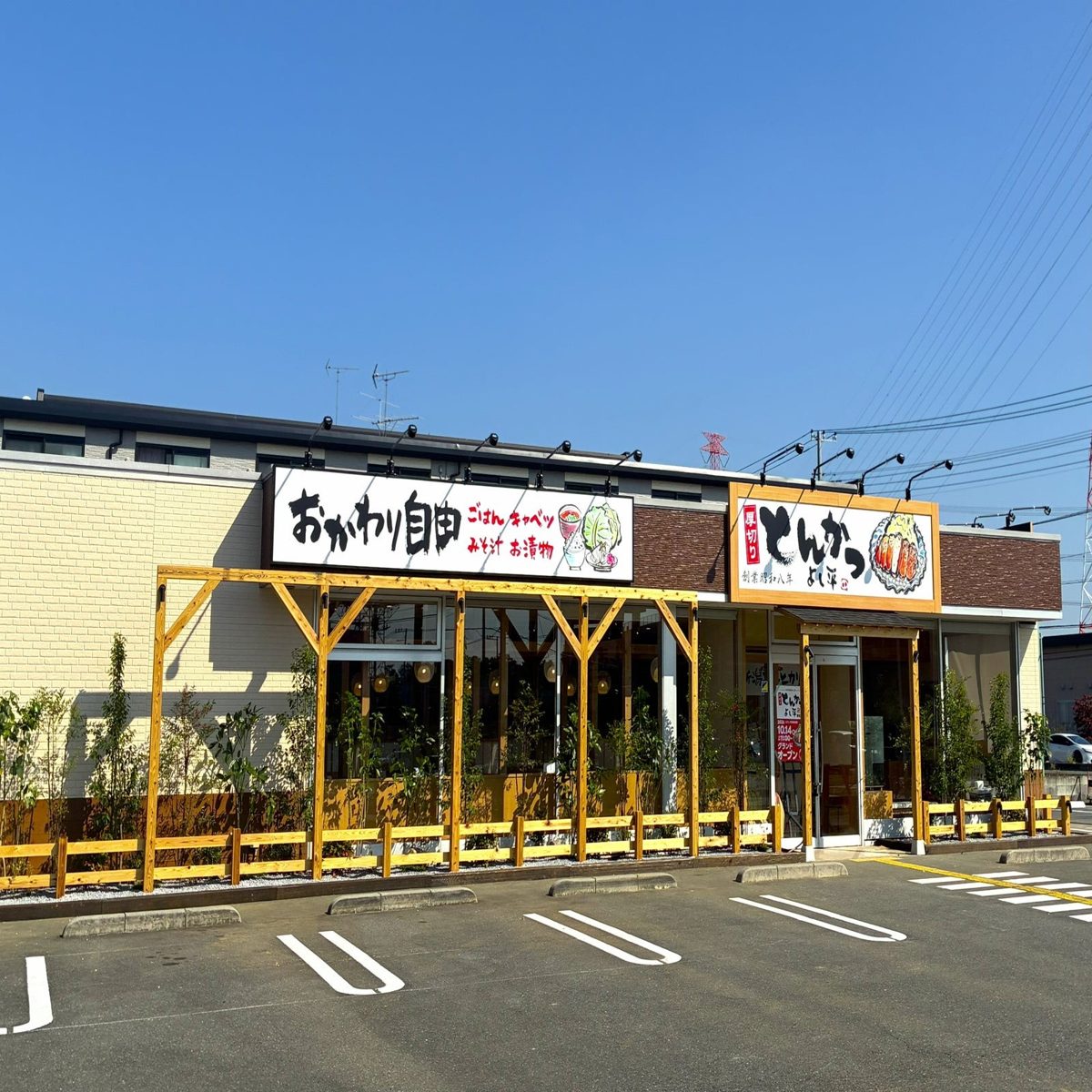黄色い外壁の平屋の店舗。「おかわり自由」の看板と「とんかつ よし平」の看板が掲げられ、手前に白線を引いた駐車場がある