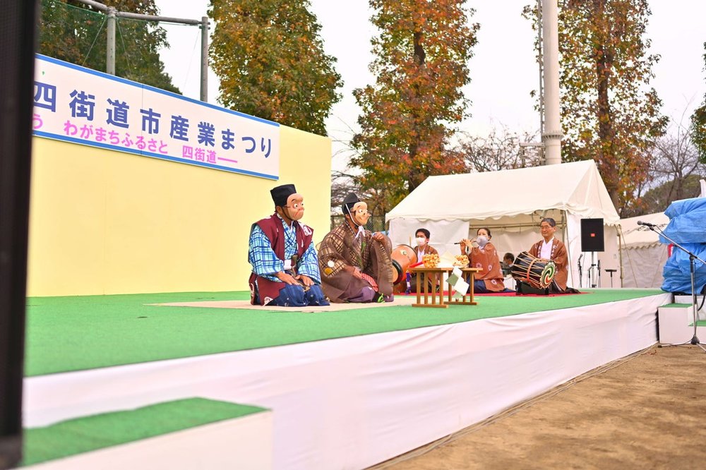 四街道市産業まつりのステージで、面をつけた演者と囃子方がお囃子を披露している様子