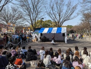 葉の落ちた並木の下、青と白のテントの前で歌う出演者を、地面に座った大勢の観客が囲んでいる