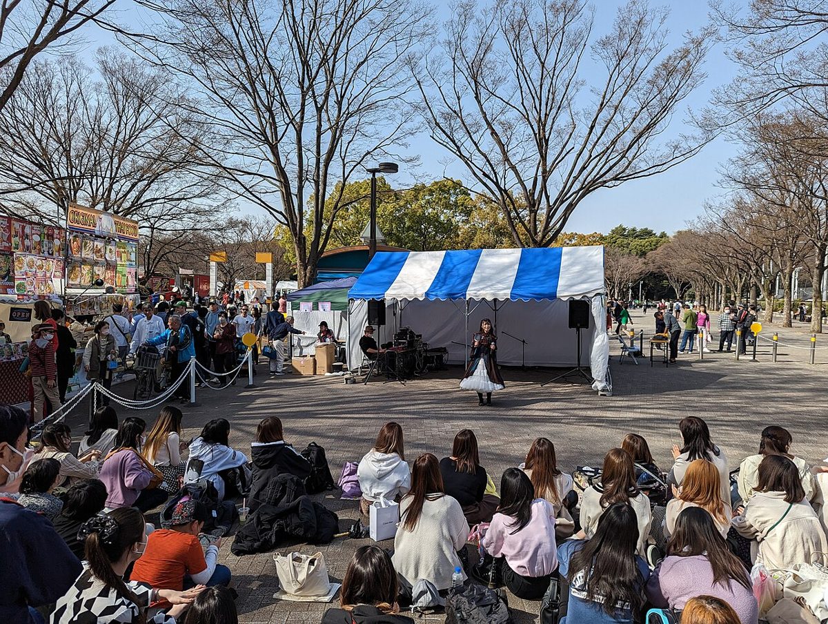 葉の落ちた並木の下、青と白のテントの前で歌う出演者を、地面に座った大勢の観客が囲んでいる